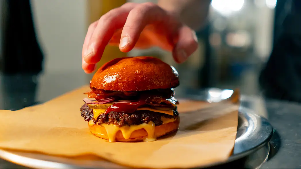 Hamburgueria com hambúrguer gourmet sendo finalizado por um chef, com pão brioche caramelizado, queijo derretido, bacon e cebola roxa.