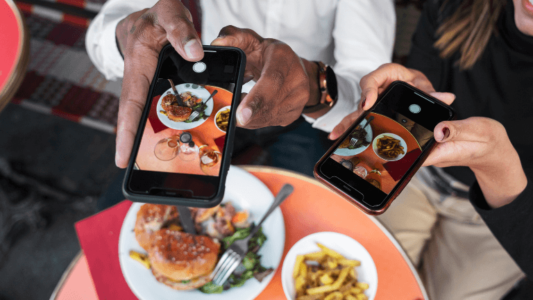 Close de mãos segurando celulares tirando fotos de pratos de comida e hambúrgueres sobre a mesa para marketing de restaurantes.